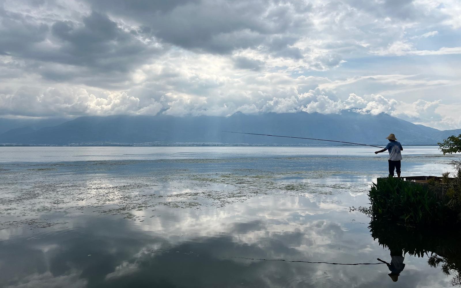 Un pescador junto a un lago tranquilo bajo un cielo de nubes.